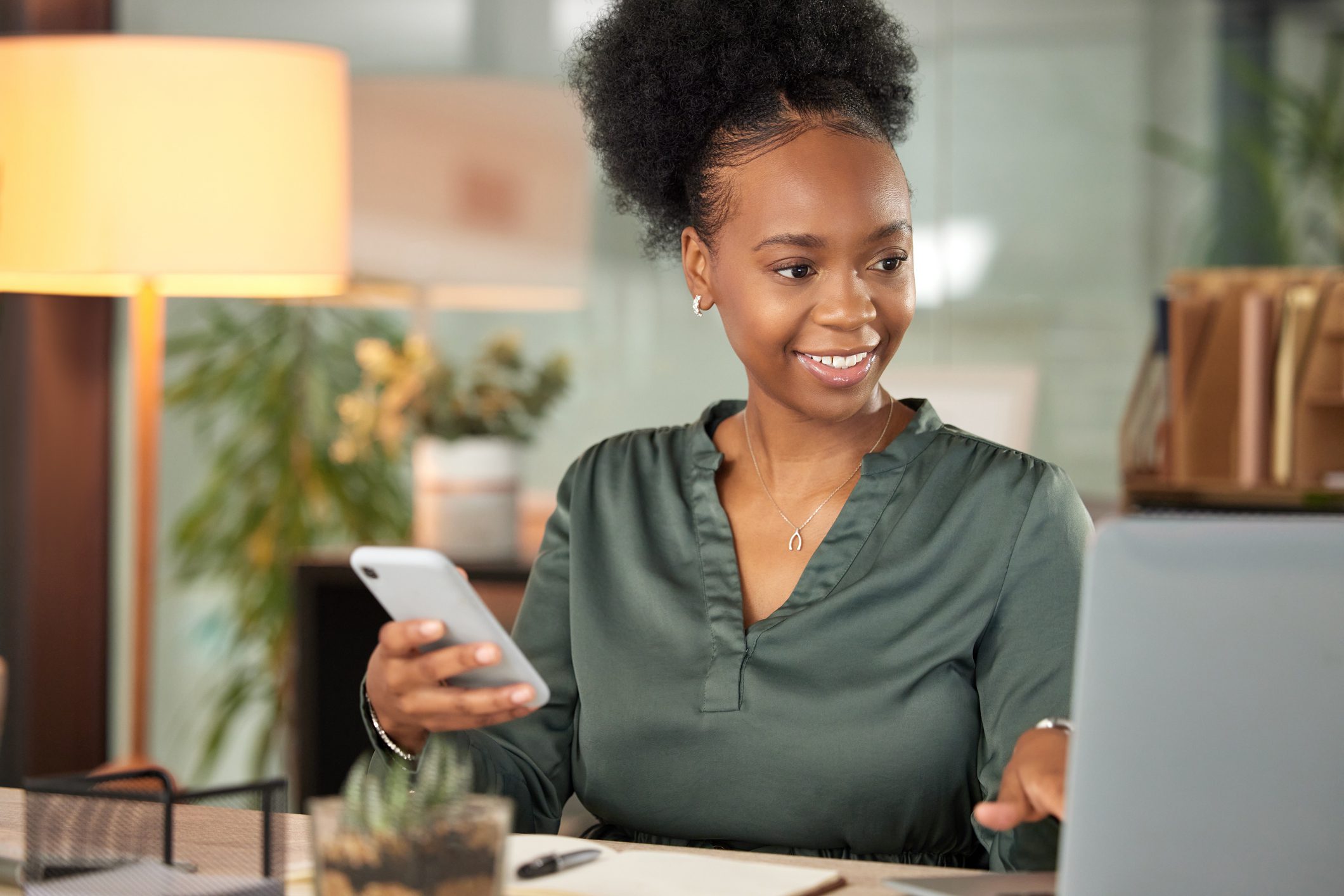 A smiling woman using a laptop and smartphone in a cozy workspace.