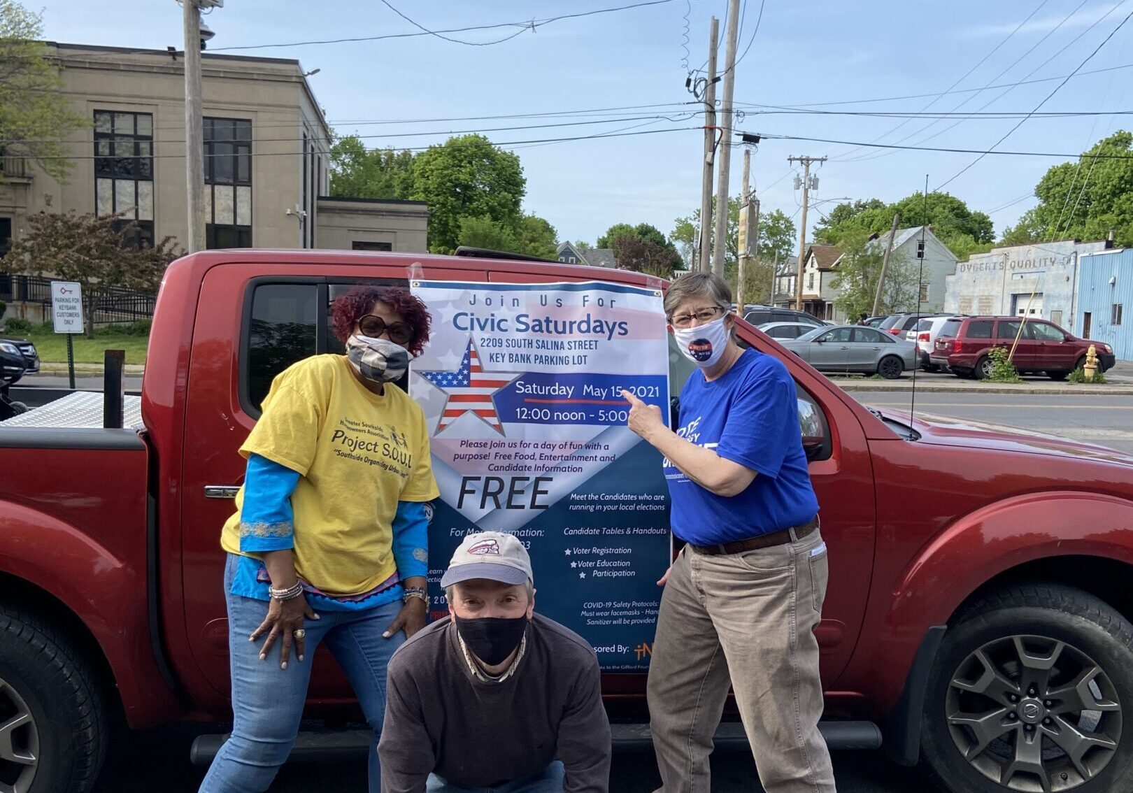Three people pose with a banner promoting an event, wearing masks outdoors.